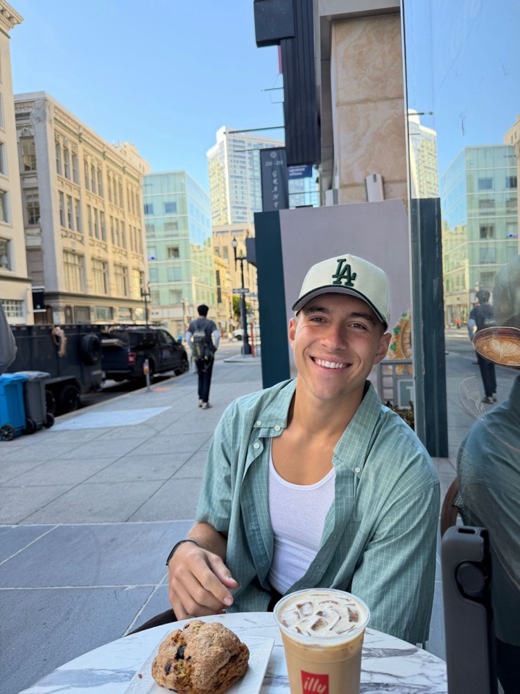Jaxson smiling at a sidewalk café table with an iced latte and a scone