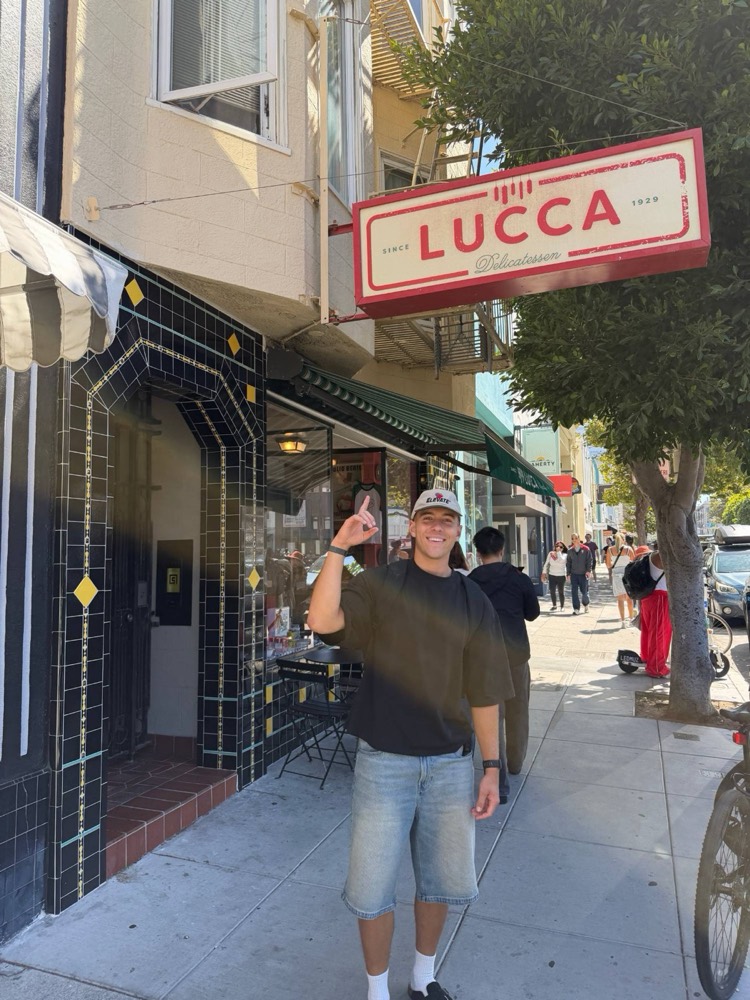 Jaxson pointing up at the Lucca Delicatessen sign on a sunny street